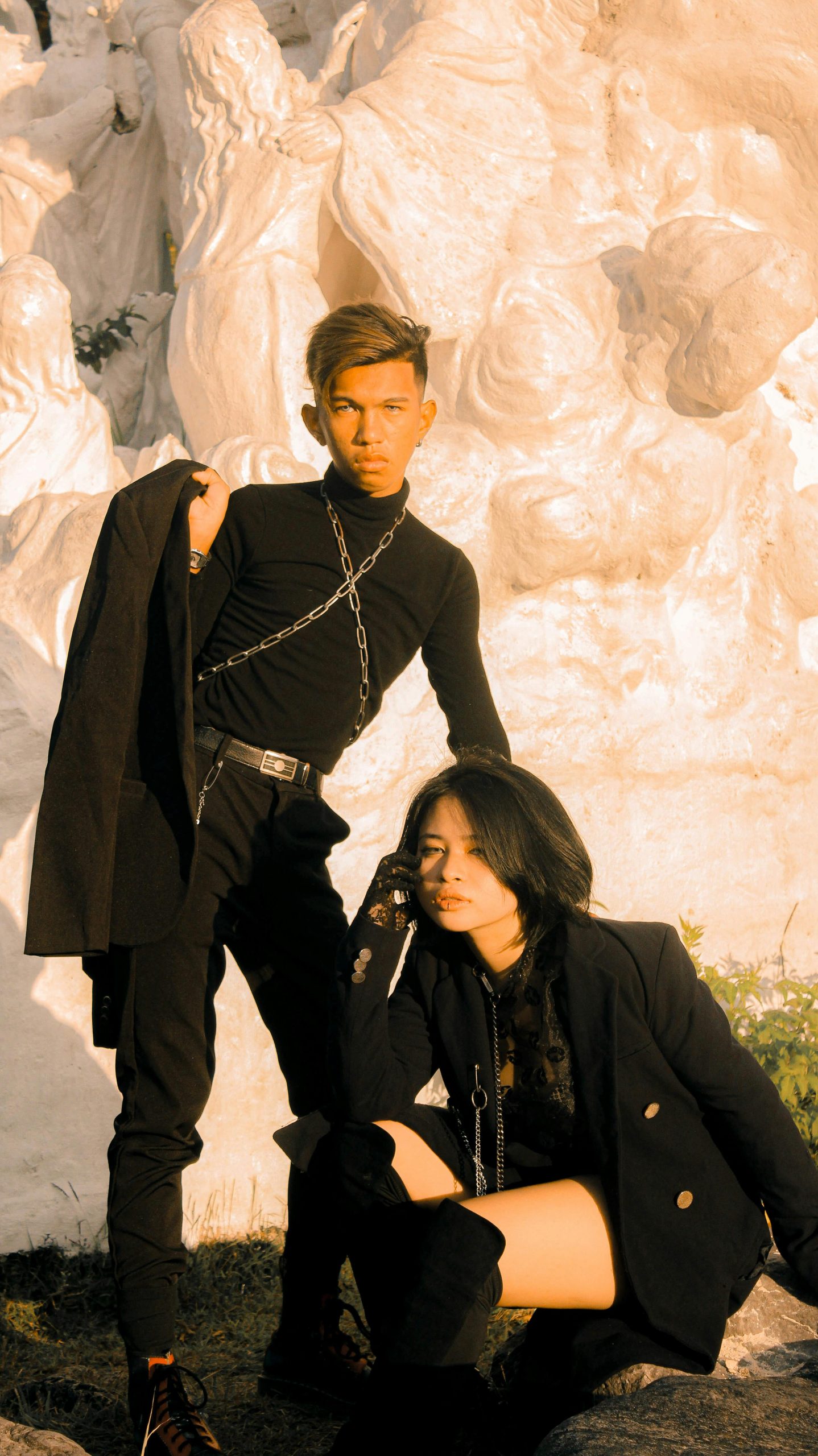 Stylish man and woman posing in fashionable clothing with a sculptural backdrop.